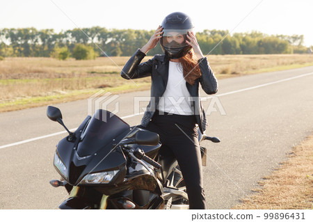 Outside shot of fast female motorcyclist wears protective helmet, poses on motorbike, stands on motorbike, covers long distance, has unforgettable journey. People, riding, safety and extreme concept Outside shot of fast female motorcyclist wears protective helmet, poses on motorbike, stands on motorbike, covers long distance, has unforgettable journey. People, riding, safety and extreme concept 99896431
