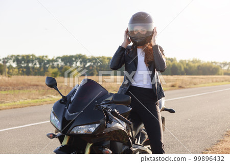 Outdoor shot of attractive confident moto driver with helmet wearing for safety, sitting on her motorbike at road, having stop, wearing leather jacket, white t shirt and black trousers. Speed concept. Outdoor shot of attractive confident moto driver with helmet wearing for safety, sitting on her motorbike at road, having stop, wearing leather jacket, white t shirt and black trousers. Speed concept. 99896432