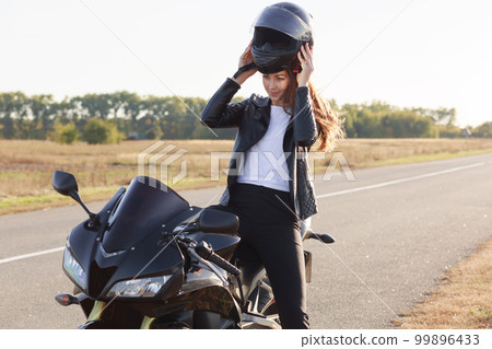 Image of beautiful slender young risk taker sitting on her motorcycle, having trip on her vehicle, fond of extreme activities, wearing helmet off, standing at side of road. Motorcycle tourism concept. Image of beautiful slender young risk taker sitting on her motorcycle, having trip on her vehicle, fond of extreme activities, wearing helmet off, standing at side of road. Motorcycle tourism concept. 99896433