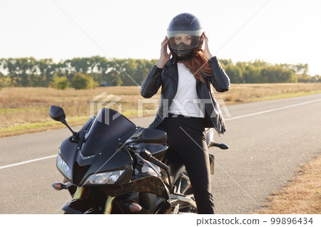 Horizontal view of stylish confident female biker dressed in fashionable clothes, sits on motorbike, wears helmet, covers long destination, poses outdoor. Motorcyclist enjoys outdoor travel. Horizontal view of stylish confident female biker dressed in fashionable clothes, sits on motorbike, wears helmet, covers long destination, poses outdoor. Motorcyclist enjoys outdoor travel. 99896434
