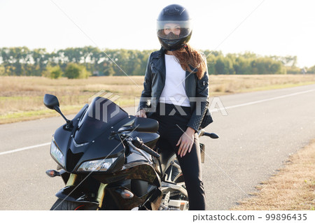 Street style concept. Active professional rider wears helmet, black leather jacket, poses on motorrbike at road, enjoys high speed, ready to cover long distance and have trip. Horizontal shot Street style concept. Active professional rider wears helmet, black leather jacket, poses on motorrbike at road, enjoys high speed, ready to cover long distance and have trip. Horizontal shot 99896435