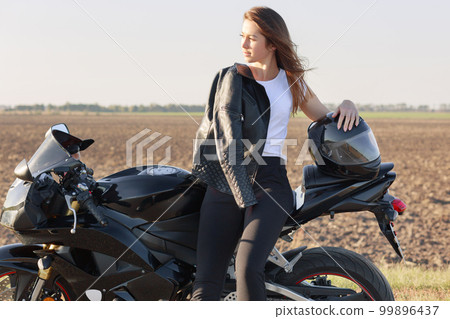 Photo of thoughtful Caucasian female likes riding motorbike, poses near her fast bike, has leather jacket on shoulder, keeps hand on helmet, looks aside with pensive expression, deep in thoughts Photo of thoughtful Caucasian female likes riding motorbike, poses near her fast bike, has leather jacket on shoulder, keeps hand on helmet, looks aside with pensive expression, deep in thoughts 99896437