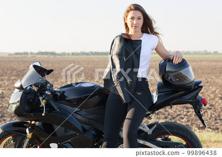 Horizontal shot of attractive motorrcyclist leans at motorrbike, keeps hand on helmet, stands near bike, has leather jacket on herself, models outdoor. People, high speed and destination concept Horizontal shot of attractive motorrcyclist leans at motorrbike, keeps hand on helmet, stands near bike, has leather jacket on herself, models outdoor. People, high speed and destination concept 99896438