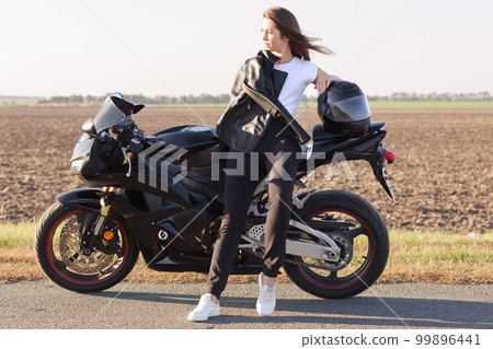 Adorable magnetic biker having trip on her motorcycle, standing near field, enjoying splendid views, looking aside, being in good mood, having confident facial expession. Extereme sports concept. 99896441