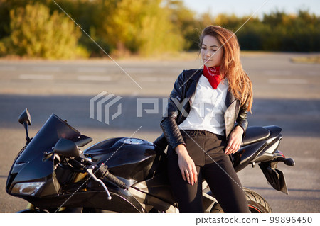 Horizontal view of thoughtful biker stands near favourite motorbike, dressed in stylish outfit, has contemplative look down, breathes fresh air, likes extreme spot. People, lifestyle and active rest 99896450