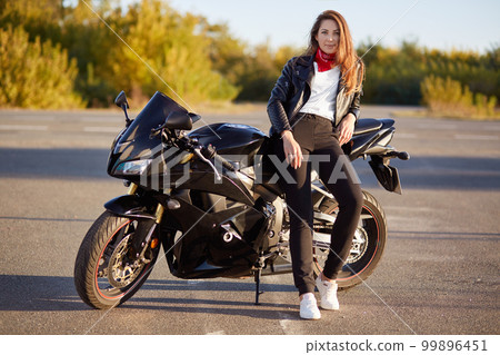 Photo of attractive woman has real adventure outdoor, stands near motorbike, dressed in bikers outfit, enjoys sunny day and calm atmosphere. People and motorcycling concept. Its time for ride 99896451