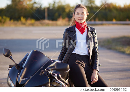 Photo of young beautiful woman stands near her cool motorrbike, wears red bandana, leather jacket, black trousers, white t shirt, looking at camera, enjoys extreme ride on bike, stop to have rest. Photo of young beautiful woman stands near her cool motorrbike, wears red bandana, leather jacket, black trousers, white t shirt, looking at camera, enjoys extreme ride on bike, stop to have rest. 99896462