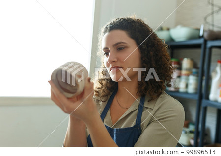 Happy concentrated woman painting clay jug with colourful paint shaping and sculpturing earthenware cup in pottery studio on ceramic workshop. Girl artist relaxing with art leisure 99896513