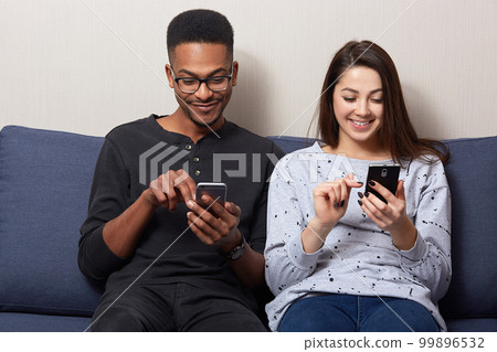Photo of young woman in casual outfit sitting on couch in room with her African boyfriend and looking at theit smartphones's on smartphone screens and loughing, see something interesting, check email. 99896532