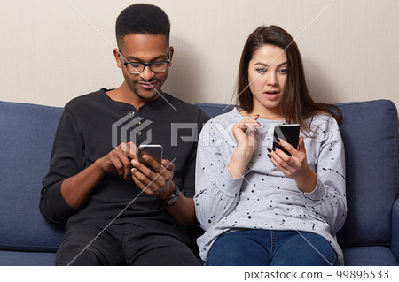 Surprised beautiful young woman holds modern cell phone, reads shocking news online, dark skinned guy wears spectacles, sits near, enjoy wireless internet connection at home, sit at comfortable sofa 99896533