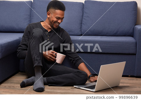 Horizontal indoorm shot of dark skinned male posing on floor near blue sofa, watches funny video on laptop computer or having video call with friend, drinks hot coffee, connected to wireless internet. Horizontal indoorm shot of dark skinned male posing on floor near blue sofa, watches funny video on laptop computer or having video call with friend, drinks hot coffee, connected to wireless internet. 99896639
