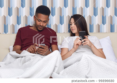 Photo of addicted mixed race lovely family couple hold modern cell phones in hands, surf social networks, sit at comfortable bed under bedclothes, connected to wireless internet, rest at home Photo of addicted mixed race lovely family couple hold modern cell phones in hands, surf social networks, sit at comfortable bed under bedclothes, connected to wireless internet, rest at home 99896680