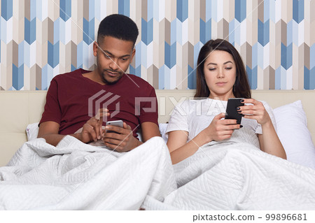 Domestic atmopshere, technology and ethnicity concept. Concentrated mixed race young woman and man use modern mobile phones, pose at bed in cozy hotel, have lazy day, chat in social networks Domestic atmopshere, technology and ethnicity concept. Concentrated mixed race young woman and man use modern mobile phones, pose at bed in cozy hotel, have lazy day, chat in social networks 99896681