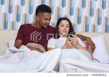 Couple in love lieing on white bed. Young darkskinned man and woman looking at mobile phone, reading or watching something interesting, handsome man hugging her wife, spending time together. Couple in love lieing on white bed. Young darkskinned man and woman looking at mobile phone, reading or watching something interesting, handsome man hugging her wife, spending time together. 99896682