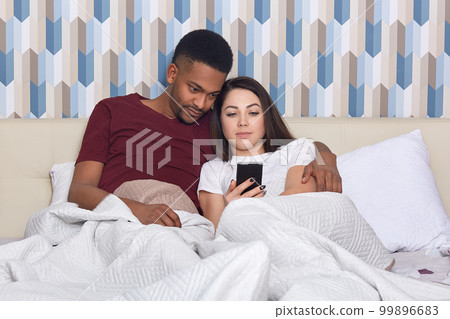 Affectionate multiethnic couple embrace together in bed, watch video or movie via cell phone, enjoy day off, high speed internet connection at home. African American guy hugs lovely woman in bedroom Affectionate multiethnic couple embrace together in bed, watch video or movie via cell phone, enjoy day off, high speed internet connection at home. African American guy hugs lovely woman in bedroom 99896683