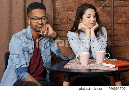Young mixed race couple have quarrel in cafeteria, displeased facial expression, sort out relationships, drink hot coffee, dont speak to each other. Unhappy multiethnic lovers in restaurant. 99896702