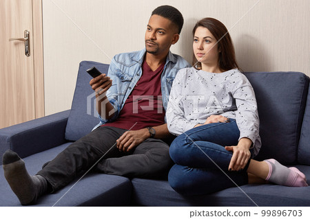 Indoor shot of multiethnic couple watch television at home on comfortable sofa. Black man holds remote control, switches on TV, sits near his young wife, have lazy day together, enjoy serial or movie 99896703