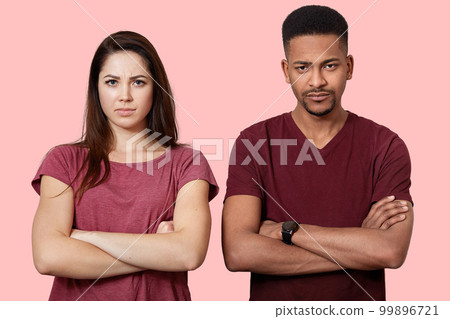 Photo of two offended young people in love, keep hands crossed, have crisis in their family, pose at pink studio wall, being displeased, look at camera, and sad. Interracial relationship concept. 99896721