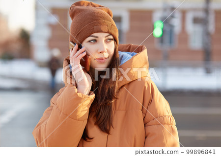 Outdoor shot of attractive Caucasian woman holds cell phone, has pleasant conversation, dressed in fashionable clothes, talks with friend or boyfriend, enjoys communication. Technology, lifestyle Outdoor shot of attractive Caucasian woman holds cell phone, has pleasant conversation, dressed in fashionable clothes, talks with friend or boyfriend, enjoys communication. Technology, lifestyle 99896841