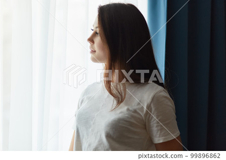 Sideways shot of thoughtful dark haired woman dressed in white t shirt, thinks about something while stands near window, enjoys calm atmosphere at home, feels lonely. Contemplative lady indoor 99896862