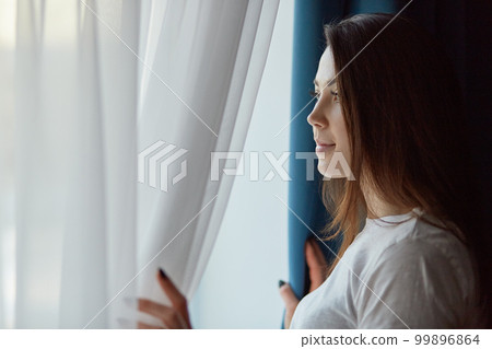 Horizontal shot of attractive brunette pensive young woman in casual white t shirt, looks out of window with thoughtful expression, has make up, enjoys calm domestic atmosphere, poses at her flat. 99896864