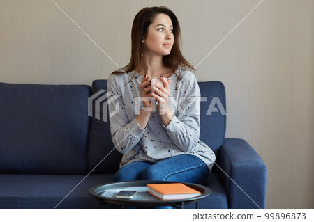 People, home concept. Thoughtful pretty European woman holds cup of hot beverage, dressed in sweater and jeans, sits at sofa, focused aside, round table with mobile phone and red notepad near 99896873
