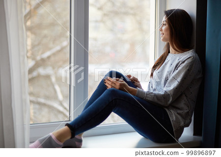 Indoor shot of thoughtful relaxed female model sits at windowsill at home, dressed in casual jumper, jeans and socks, admires beautiful view during winter from window, has weekend or day off Indoor shot of thoughtful relaxed female model sits at windowsill at home, dressed in casual jumper, jeans and socks, admires beautiful view during winter from window, has weekend or day off 99896897