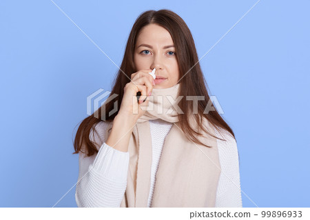 Young dark haired woman wearing white sweater and scarf standing dripping nasal drops isolated on blue background, ill lady looks at camera with upset facial expression. Young dark haired woman wearing white sweater and scarf standing dripping nasal drops isolated on blue background, ill lady looks at camera with upset facial expression. 99896933