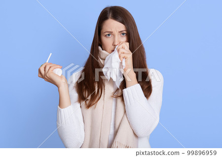 Dark haired unhealthy Caucasian woman with napkin sneezing and holding throat spray in hands to help herself. Rhinitis, cold, sickness and allergy, female uses handkerchief, wearing casual attire. 99896959