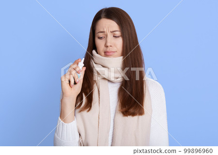 Young sad woman posing isolated over blue background, holding nasal spray in hands and looking on it with upset expression, being infected corona virus, don't want using spray. 99896960