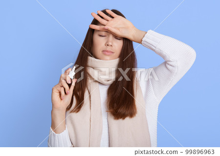 Sick female wearing sweater and wrapped in scarf, holding nasal spray in hand, touching her forehead, keeping eyes closed, suffering from fever, being infected with covid19. 99896963