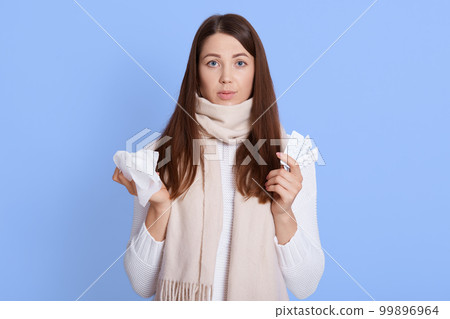 Portrait of cute unhealthy brown haired female in white sweater and scarf, holding napkin for blowing nose, looking at camera, desperate female has flu or corona virus. 99896964
