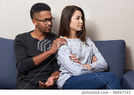 Offended attractive woman sitting back to her husband on sofa in flat, african male touches girl's shoulders and asks to forget him, couple wearing casual outfit. Quarrel and relationship concept. Offended attractive woman sitting back to her husband on sofa in flat, african male touches girl's shoulders and asks to forget him, couple wearing casual outfit. Quarrel and relationship concept. 99897145