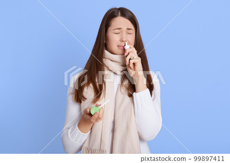 Caucasian female frowning face while using nasal spray, holds throat spray in hands, keeps eyes closed, dark haired sick girl dresses jumper and scarf being ill, needs treatment, against blue wall. Caucasian female frowning face while using nasal spray, holds throat spray in hands, keeps eyes closed, dark haired sick girl dresses jumper and scarf being ill, needs treatment, against blue wall. 99897411