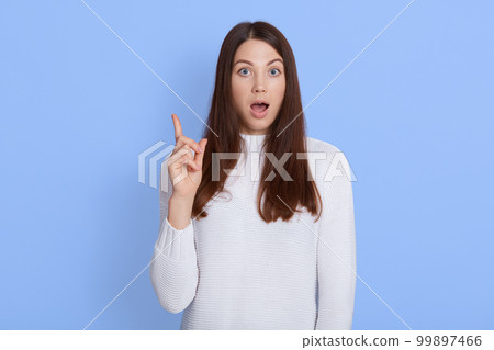 Amazed shocked young woman pointing up with fore finger, keeps mouth widely opened, wearing casual white shirt, has dark hair, being shocked and astonished, stands against blue wall. 99897466