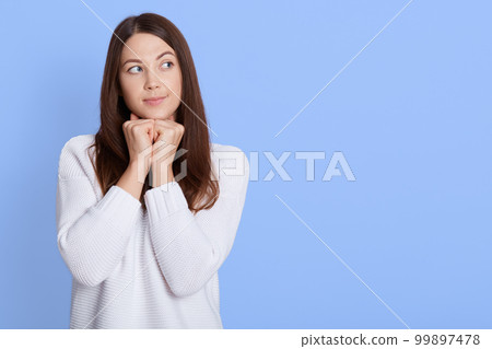Good looking thoughtful young European female keeping hand under chin, has healthy skin, wears white sweater, looks pensive aside, contemplates about something, poses over blue wall. Copy space. Good looking thoughtful young European female keeping hand under chin, has healthy skin, wears white sweater, looks pensive aside, contemplates about something, poses over blue wall. Copy space. 99897478