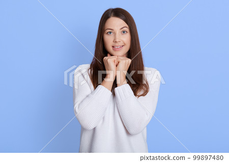 Portrait of charming girlfriend with dark hair, holding fists under chin and smiling broadly while staring focused at camera, waiting for something beautiful, posing over blue wall. 99897480