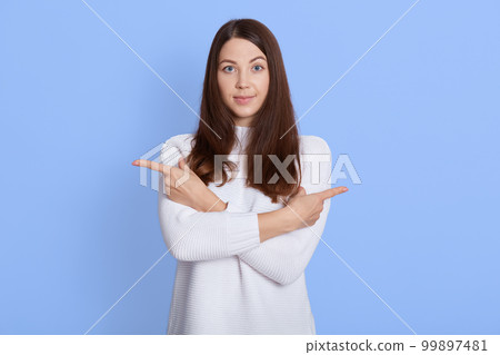 Young beautiful woman wearing casual white shirt over blue background, pointing to both sides with fingers, different direction disagree, looks at camera, choosing where to go. Young beautiful woman wearing casual white shirt over blue background, pointing to both sides with fingers, different direction disagree, looks at camera, choosing where to go. 99897481