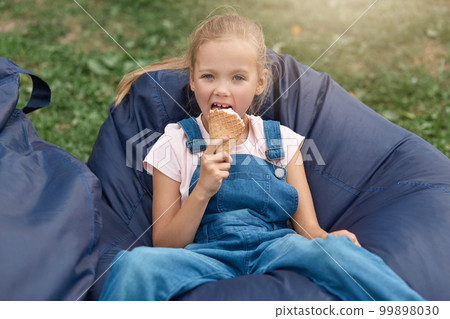 Happy little child, adorable toddler girl with blond hair and ponytail,wearing casual outfit, enjoying ice cream outdoors while sitting on blue frameless chair in amusement park on sunny summer day 99898030