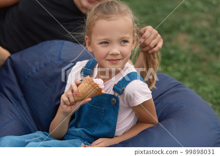 Adorable kid girl holding ice cream in hands, sitting next to fathe on farameless chair outdoor, father and his little doughter spending time outdoor together. Family, lifestyle and happyness concept. 99898031