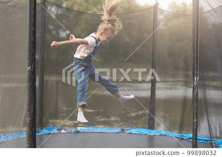 Full length photo of happy girl jumping high on trampoline in park, blonde female chicd with ponytail wearing white shirt and denim overalls, kid spending time in open air, having fun outdoor. 99898032