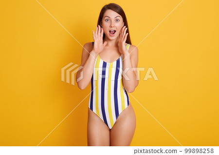 Photo of shocked young woman in srtiped swimsuit looking directly at camera, isolated over yellow studio wall, keeps hands near mouth, crying something, posing with widely opened eyes. People concept. 99898258
