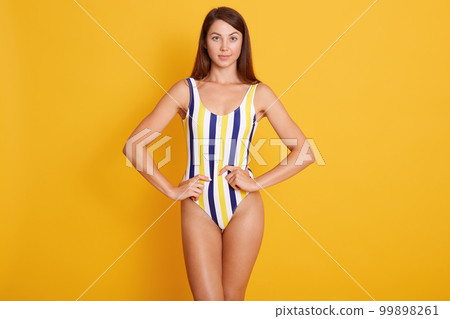 Fashion photo of beautiful and young lady with pefect body asnd skin, wearing in striped swimsuits posing in studio isolated over yellow wall, looks at camera, model has pleasant appearance. 99898261