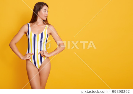 Photo of young woman with straight dark hair dressed in swimsuit, looking aside isolated over yellow background in studio, keeps hjands on hips, copy space for advertismant or promotion text. Photo of young woman with straight dark hair dressed in swimsuit, looking aside isolated over yellow background in studio, keeps hjands on hips, copy space for advertismant or promotion text. 99898262