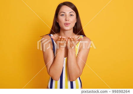 Horizontal shot of pleasant looking female with healthy skin, wearing swimsuit, making air kiss gesture, flirts with handsome guy at seaside, isolated over yellow studio background. People concept. 99898264