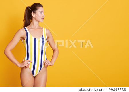 Picture of young woman dresses stylish striped swimwear isolated over yellow background, adorable female looking aside and keeping hands on hip. Copy space for advertisment or promotion text. Picture of young woman dresses stylish striped swimwear isolated over yellow background, adorable female looking aside and keeping hands on hip. Copy space for advertisment or promotion text. 99898288