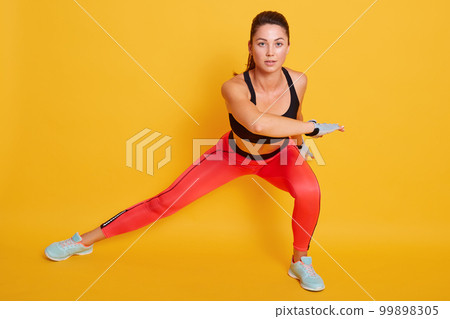 Studio shot of young beautiful woman working out, doing side lunges, slimledy exercises for legs, hips and buttocks, sporty female wears black top and leggins looks at camera, isolated on yellow wall. Studio shot of young beautiful woman working out, doing side lunges, slimledy exercises for legs, hips and buttocks, sporty female wears black top and leggins looks at camera, isolated on yellow wall. 99898305