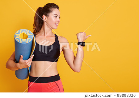 Powerful strong Caucasian woman raises arm, shows biceps, holding blue fitness mat for gym training, dresses sportwear, isolated over yellow background, lady ready for workout or yoga practice. 99898326