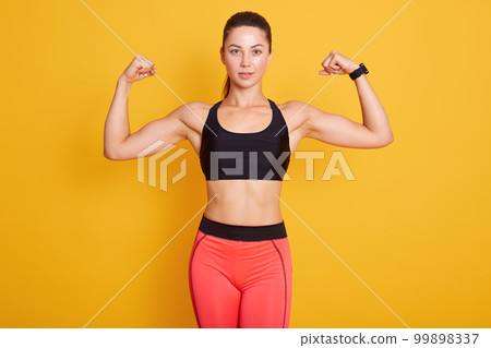 Close up portrait of young cute sporty brunette woman with ponitail, showing her arms and biceps to camera, female wears black bra and leggins. Sport, healthy lifestyle, women health, fitness concepts Close up portrait of young cute sporty brunette woman with ponitail, showing her arms and biceps to camera, female wears black bra and leggins. Sport, healthy lifestyle, women health, fitness concepts 99898337