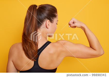 Horizontal shot of attractive fitness woman posing backwards isolated over yellow studio background. Trained female body, girl showing her biceps, lady with ponitail wearing black sport top. Horizontal shot of attractive fitness woman posing backwards isolated over yellow studio background. Trained female body, girl showing her biceps, lady with ponitail wearing black sport top. 99898341